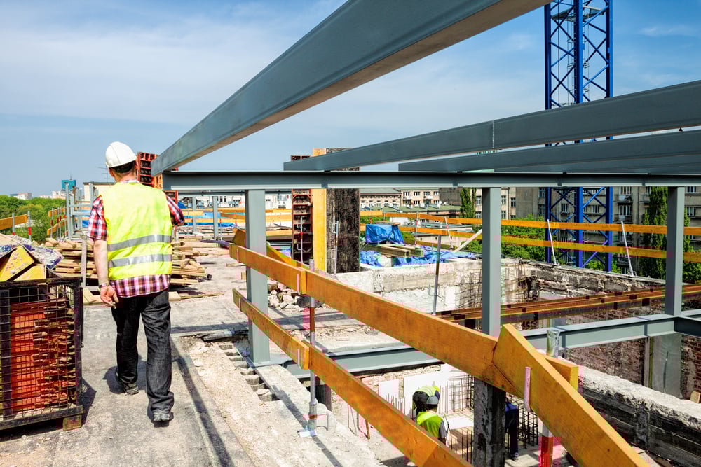 A worker in a hard hat and vest tours the construction of a new, large building, where steel beams rise from the foundation.